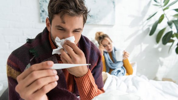 sick man with fever holding thermometer and napkin in bedroom with woman behind